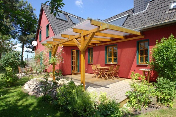 A red house with a wooden pergola, a patio with a table and chairs, and a garden with rocks and plants.