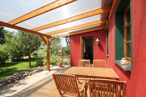 Puinen terassi, jossa on pöytä ja tuoleja, pergola, jonka varassa kasvaa köynnöskasveja, sekä punainen rakennus, jossa on ikkuna ja ovi.