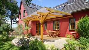 Una casa rossa con una pergola in legno, un patio con tavolo e sedie, e un giardino con rocce e piante.