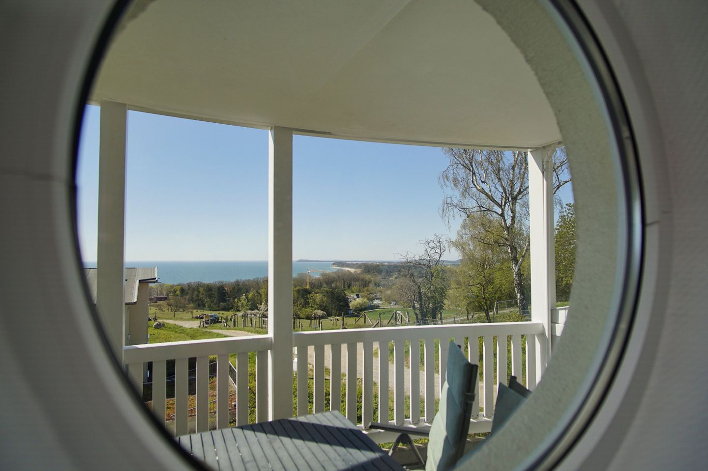 A view through a circular window of a porch with a white railing, overlooking a landscape with trees and a distant body of water.