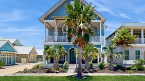 A two-story house with a balcony, surrounded by palm trees and a well-maintained lawn. - Chic Sunset Views on Palmilla Golf Course, Designer Interior & Chef’s Kitchen (Port Aransas)