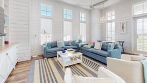 A spacious living room with a large sectional sofa, a coffee table, and a view of the outdoors through large windows. - Chic Sunset Views on Palmilla Golf Course, Designer Interior & Chef’s Kitchen (Port Aransas)