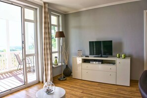 A room with a TV on a cabinet, a vase, and a table with a glass object. - With sea view and balcony, use of swimming pool and sauna - Sea view apartment 22 - (Göhren)
