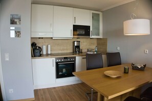 A compact kitchen with white cabinets, a microwave, a coffee maker, and an oven. There is a dining table with chairs and a basket on the table.
