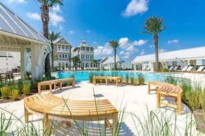 Zona de piscina con bancos de madera, palmeras, una piscina y un edificio con balcones.