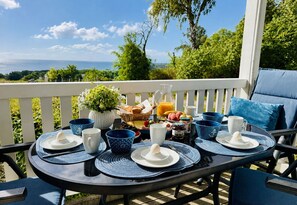 Une table dressée sur un balcon avec vue sur l’océan, où est présenté un assortiment de produits pour le petit-déjeuner, comprenant des fruits, du pain et des boissons.