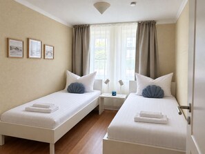 Une chambre d’hôtel avec deux lits simples, une petite table avec deux lampes, une fenêtre avec des rideaux et des tableaux encadrés au mur.