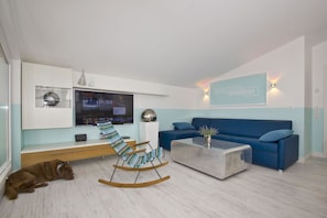 A modern living room with a blue sofa, a wooden rocking chair, a coffee table, and a TV mounted on the wall. - With a view of the sea and a balcony, access to the pool and sauna - Sea View Apartment 67 - (Göhren)