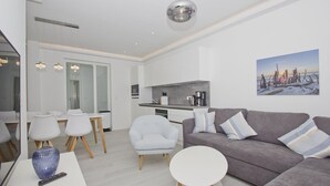 A modern living room with a grey sofa, white armchairs, and a round coffee table. A kitchen with a grey countertop and a coffee machine is visible in the background.