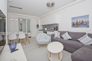 A modern living room with a grey sofa, white armchairs, and a round coffee table. A kitchen with a grey countertop and a coffee machine is visible in the background. - Modern vacation apartment with private terrace - Haus Inselwind FeWo MEERsalz - House (Gager)