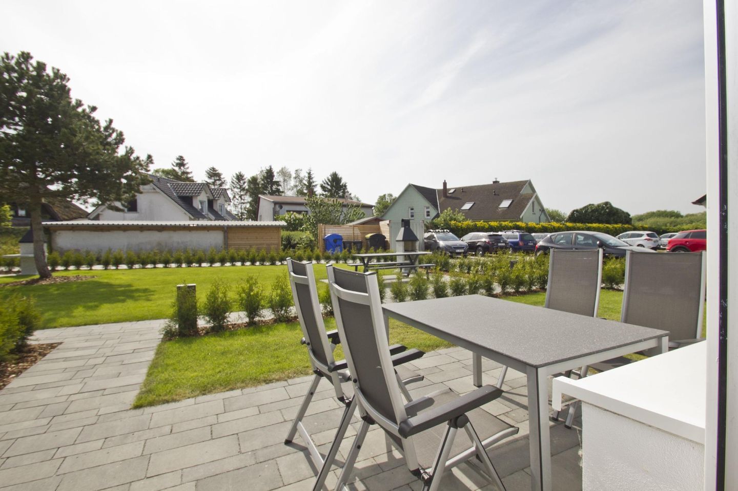 Een buitenpatio met een tafel en stoelen, een pad en zicht op geparkeerde auto's en huizen op de achtergrond.