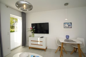 A modern living room with a flat-screen TV, a white sofa, a dining table with chairs, and a decorative vase. - Modern vacation apartment with a private terrace - Haus Inselwind FeWo MEERstille - (Gager)