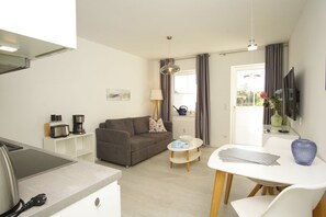 A compact living space with a kitchenette, a grey sofa, a small round table, and a TV mounted on the wall.