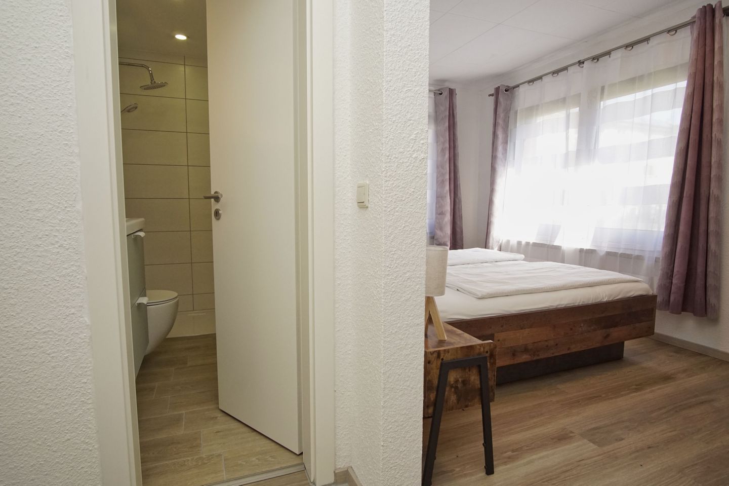 A bedroom with a wooden bed, a small wooden stool, a window with curtains, and a bathroom with a shower and toilet.