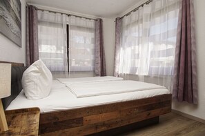 Un dormitorio con cama de madera, ropa de cama blanca, cortinas traslúcidas y vistas al exterior.