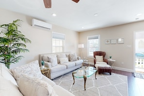 A living room with a sofa, a glass coffee table, a side table, and a chair. There is a potted plant, a lamp, and a window with blinds.