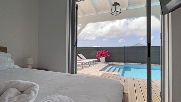 A bedroom with a large bed, a view of a swimming pool, and a balcony with lounge chairs and a flower pot. - Architect designed villa for 8 with swimming pool (Saint François)