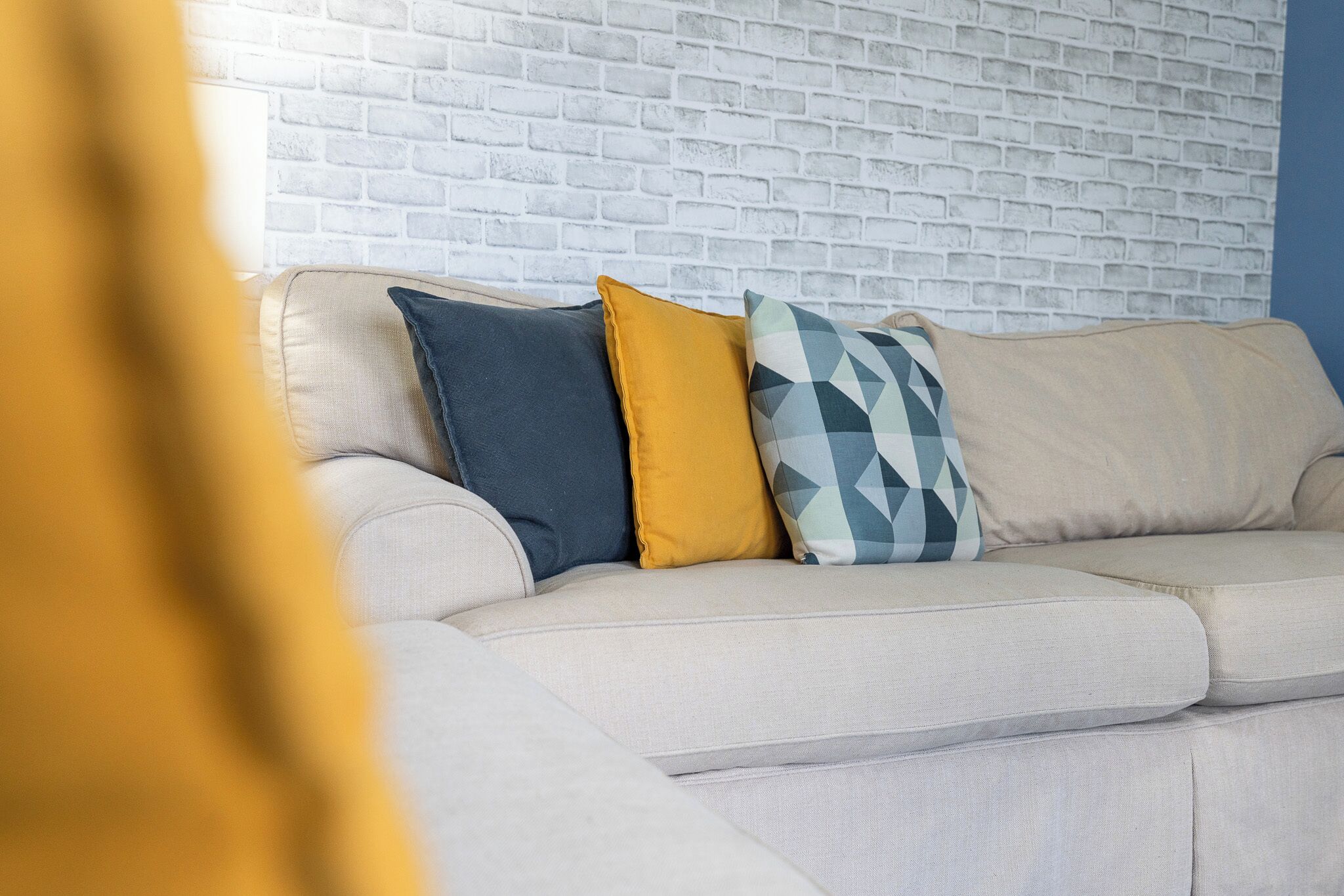 A beige sofa with three cushions against a white brick wall.