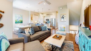 A modern living room with a grey sofa, a coffee table, and a television. There is a kitchen in the background with a sign that reads "WINERY". - Luxe Palmilla townhome with resort pool & golf - walk to beach (Port Aransas)