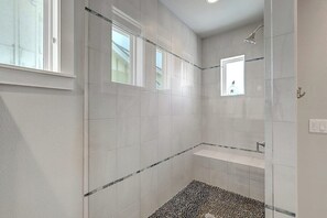 Ein Badezimmer mit weißen Fliesen, einer Dusche, einem Fenster und einem kleinen Ablagebrett.