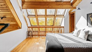 A bedroom with a sloped ceiling, wooden beams, a large window with a view of autumn trees, a bed with a quilt, and a small stool.
