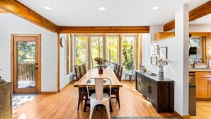 A spacious dining area with a wooden table and chairs, large windows with a view of trees, and a kitchen with wooden cabinets.