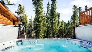 A hot tub surrounded by tall pine trees, with a wooden deck and a satellite dish on the roof.