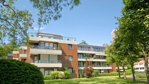 Un immeuble résidentiel à plusieurs étages, doté de balcons, d’une pelouse bien entretenue et entouré d’arbres.