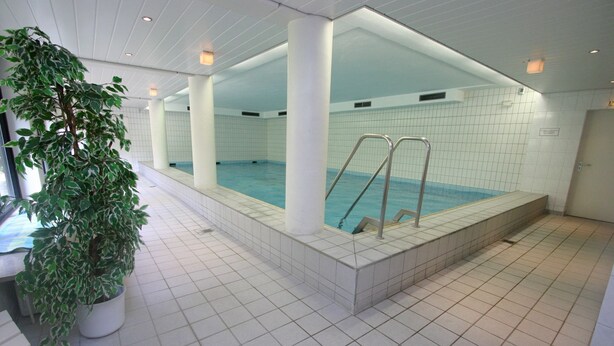 Une piscine dont le sol et les murs sont recouverts de carreaux, entourée de colonnes blanches, et avec un pot de fleurs placé sur la gauche.
