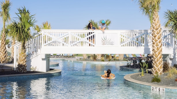 Un espace piscine agrémenté d’un pont blanc, de palmiers et d’une personne sur un flotteur.
