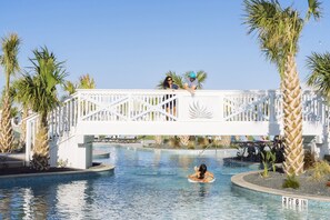 A pool area with a white bridge, palm trees, and a person in a float. - Beach Resort, Pool and Golf Course Living at Palmilla Beach - Port Aransas, TX (Port Aransas)