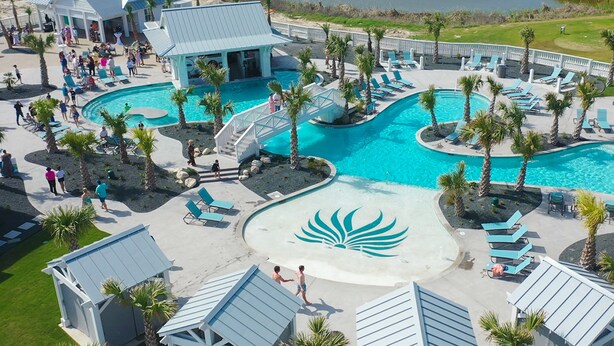 Et resort med en stor central pool, palmer og en lille bygning med blå tag.
