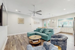 A modern living room with a sectional sofa, a coffee table, patterned armchairs, a ceiling fan, and a kitchen area with a window.