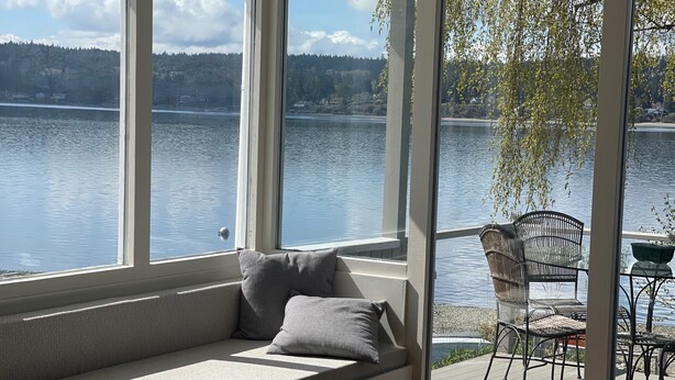 A room with a large window overlooking a lake, a seating area with cushions, and a patio with outdoor furniture. - Prime Sandy Beachfront Home with Mountain Views - 5 minutes to Gig Harbor/Tacoma (Gig Harbor)