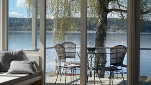 A room with a large window overlooking a lake, furnished with wicker chairs and a table. - Prime Sandy Beachfront Home with Mountain Views - 5 minutes to Gig Harbor/Tacoma (Gig Harbor)
