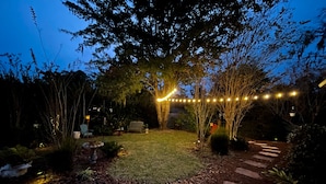 Un sentier de jardin, ponctué de pierres plates, mène à un banc placé sous un arbre, le tout étant entouré de plantes et illuminé par des guirlandes lumineuses.