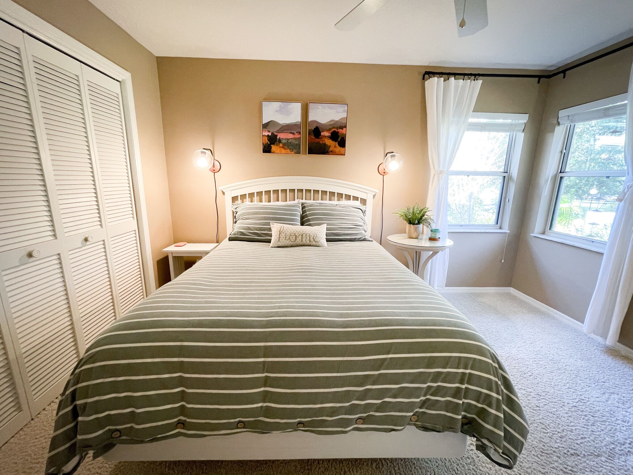 A bedroom with a bed, two wall-mounted lamps, a small round table with a plant, and a window with curtains.