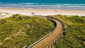 Ein hölzerner Gehweg schlängelt sich durch eine Wiese mit Strand und Meer im Hintergrund.