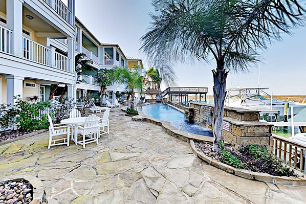 A pool area with stone flooring, white lounge chairs, and a palm tree. - Coastal retreat with pool, balconies, sundeck & boat slip - near beach (Corpus Christi)