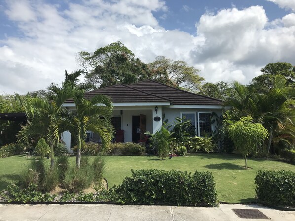 A single-story house with a dark tiled roof, surrounded by lush greenery and a well-maintained lawn.
