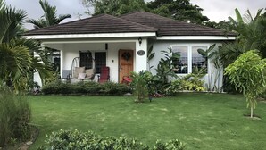 A single-story house with a tiled roof, a front porch with a swing chair, and a well-maintained lawn. - Sweet Oasis - gated with 24/hr security/ swimming pool/ free access to beach  (Saint Ann's Bay)