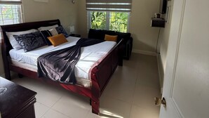 A bedroom with a wooden bed, a ceiling fan, a window with blinds, and a small shelf with a plant. - Sweet Oasis - gated with 24/hr security/ swimming pool/ free access to beach  (Saint Ann's Bay)