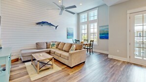 A spacious living room with a sectional sofa, wooden flooring, a ceiling fan, and large windows. - Golf-View Palmilla Beach retreat with pool, private garage, & alfresco dining (Port Aransas)