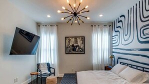 Een slaapkamer met een bed, een stoel, een bankje, een televisie en een schilderij aan de muur.