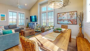 A spacious living room with a large wooden dining table, comfortable seating, a television, and large windows. - Palmilla Beach gem - 2 luxe suites, 2 balconies, pools, golf-course views & w/d (Port Aransas)
