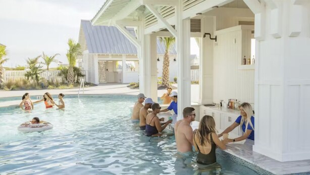 A pool area with people swimming and socializing, a bar area with a counter, and a covered seating area with a view of the pool. - Palmilla Beach gem - 2 luxe suites, 2 balconies, pools, golf-course views & w/d (Port Aransas)