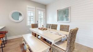 A dining area with a long table, wicker chairs, and a round mirror on the wall.