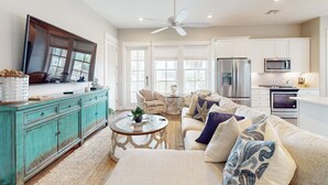 A spacious living room with a large TV, a teal cabinet, a beige sofa with patterned cushions, and a kitchen area with modern appliances.