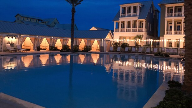 A swimming pool with a reflection of buildings and palm trees, surrounded by a covered area with string lights. - Gulf-View Palmilla Resort Condo | Pool, On-Site Dining & Beach Boardwalk (Port Aransas)