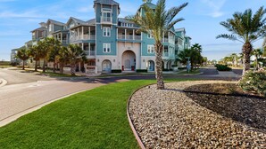 A large, multi-story building with a light blue facade, multiple balconies, and a well-maintained lawn. - Gulf-View Palmilla Resort Condo | Pool, On-Site Dining & Beach Boardwalk (Port Aransas)